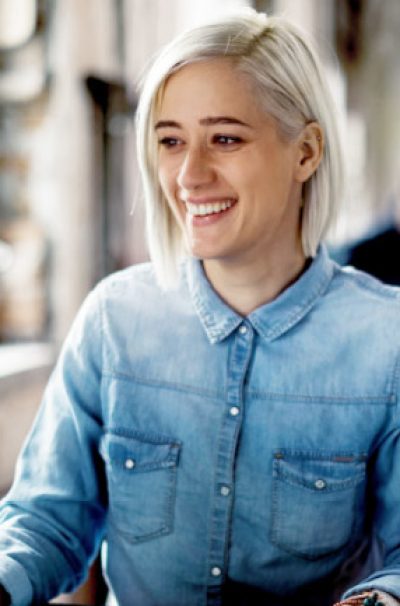 Junge blonde Frau, die an einem Tisch vor einem Monitor sitzt und lächelt